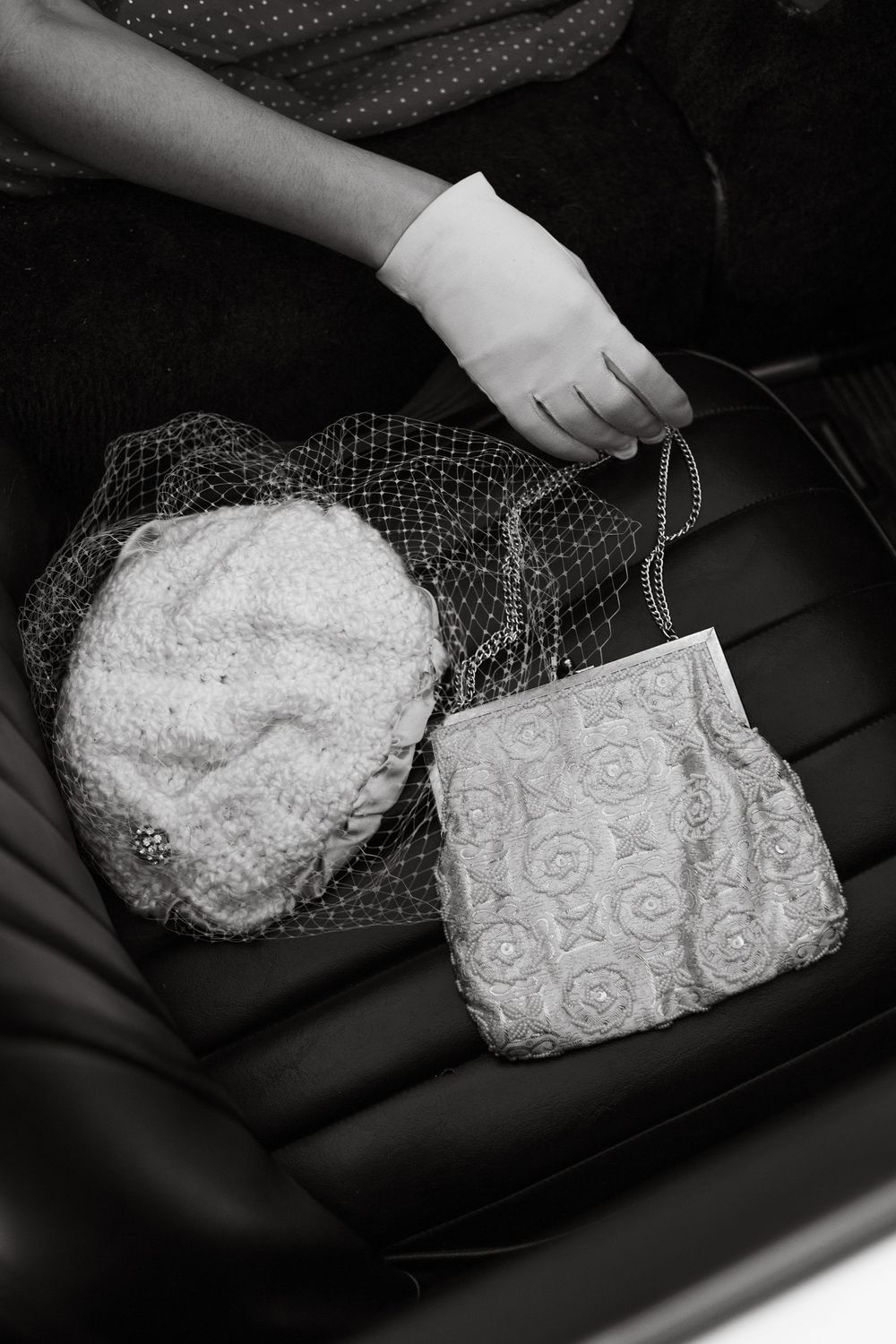 Black and white photo of a gloved hand holding a floral beaded purse beside a lace hat with veil on a car seat.