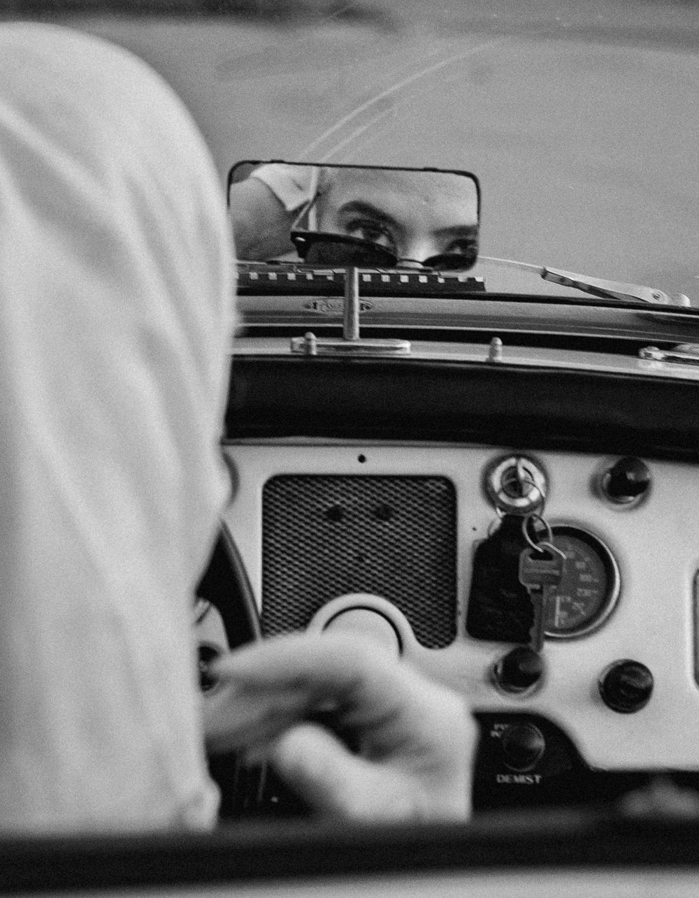 Black and white photo of a driver reflected in a rearview mirror inside a vintage vehicle dashboard.