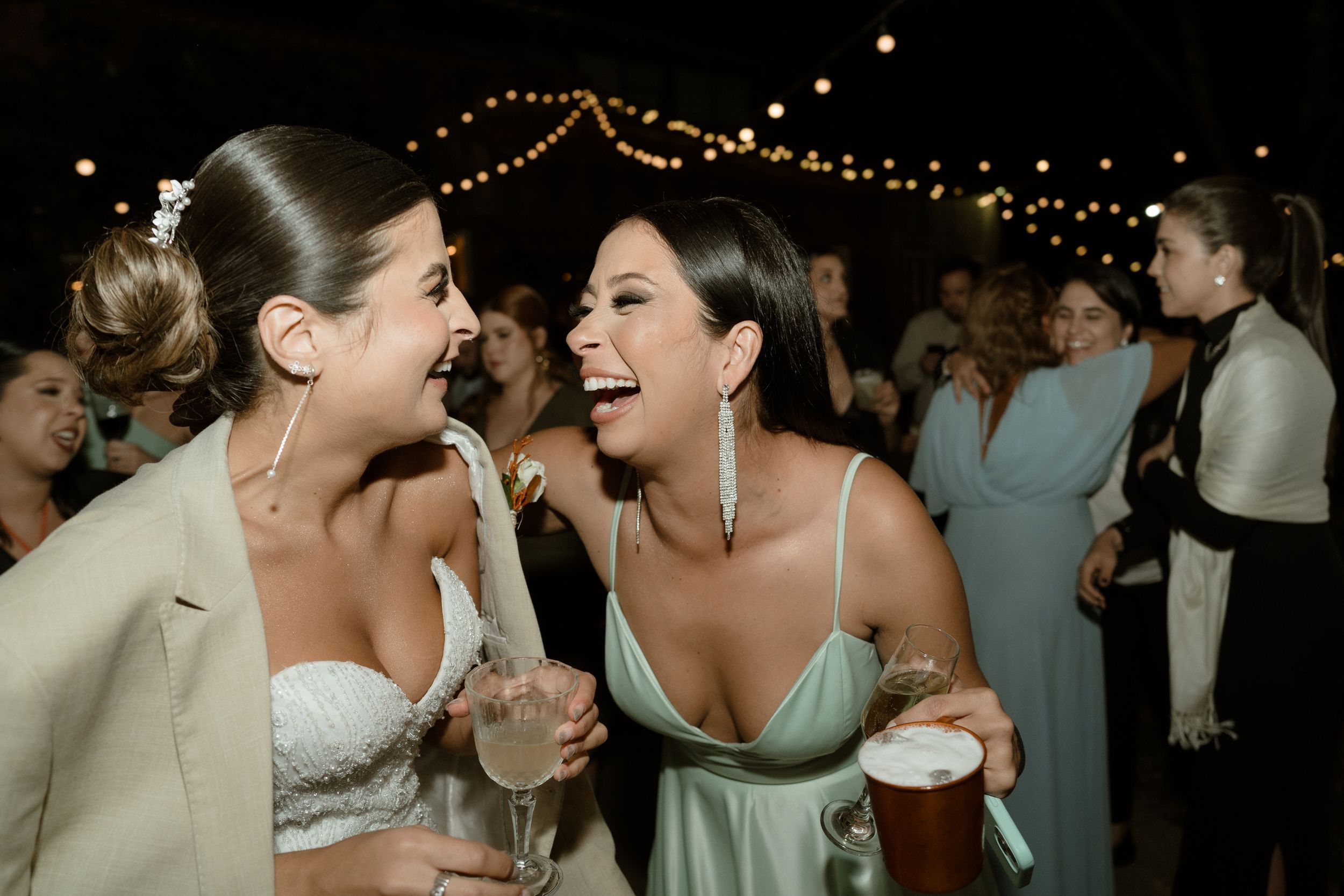 Two friends sharing laughter during a celebration while holding drinks outdoors at night.