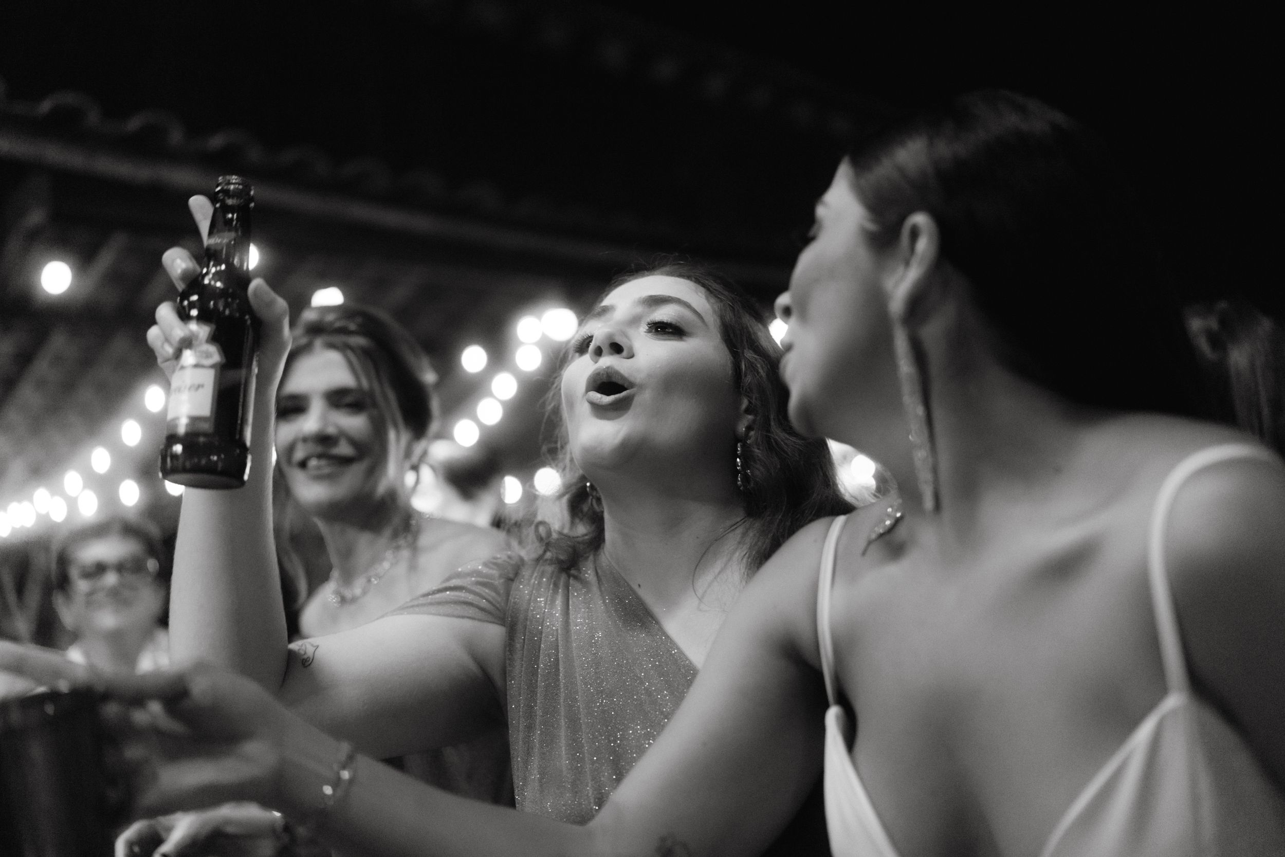 Three wedding guests celebrating with drinks under twinkling lights at an outdoor reception.