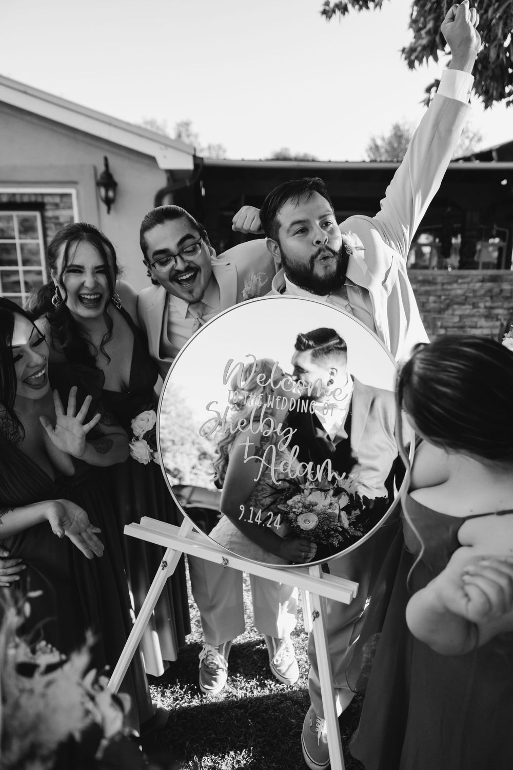 Wedding guests laugh while looking at a reflective mirrored wedding sign display outdoors.