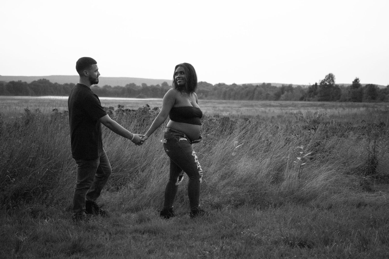 A couple holds hands while standing together in a grassy field at sunset in black and white.