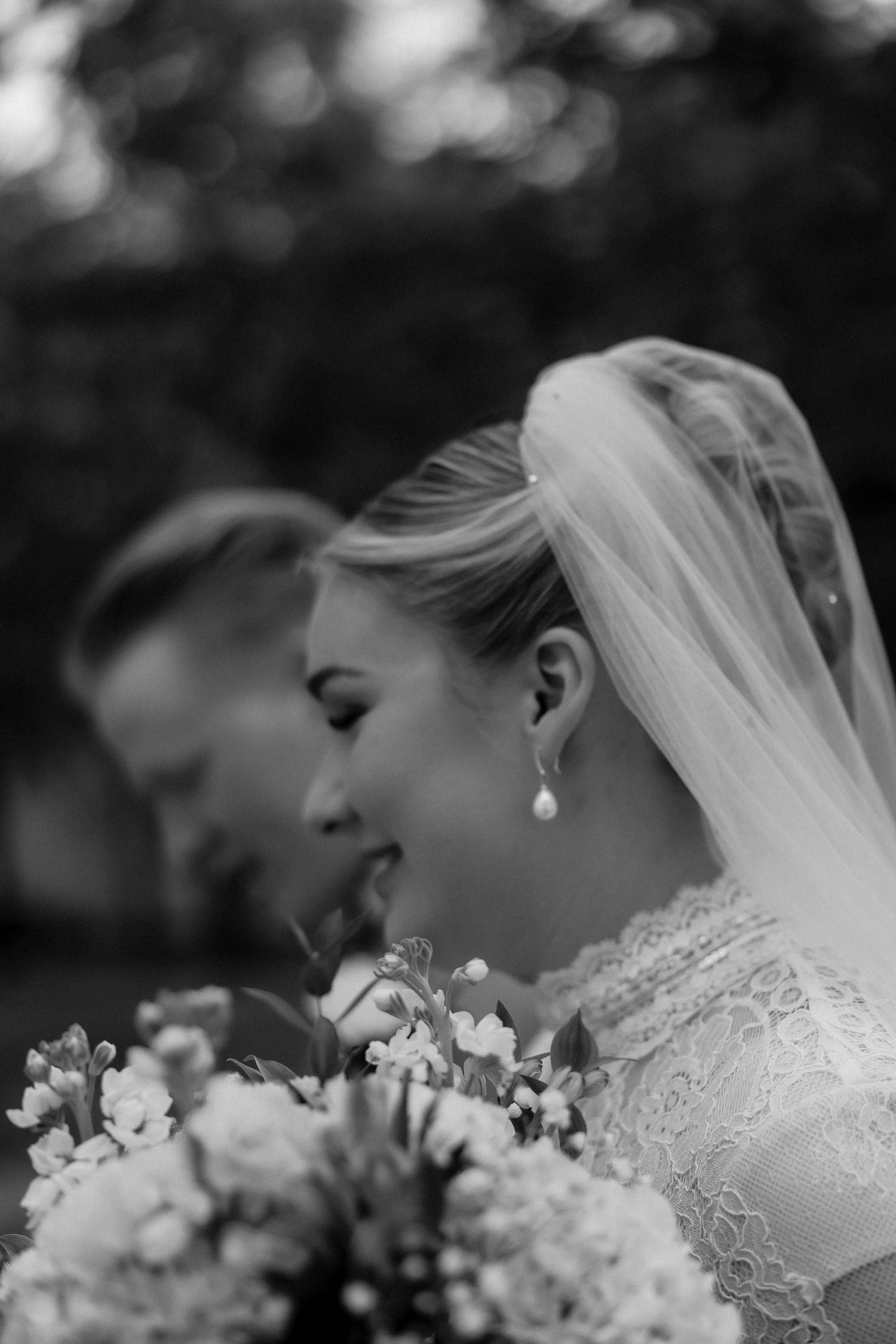 Side profile of bride in lace dress and veil holding bouquet with soft bokeh background.