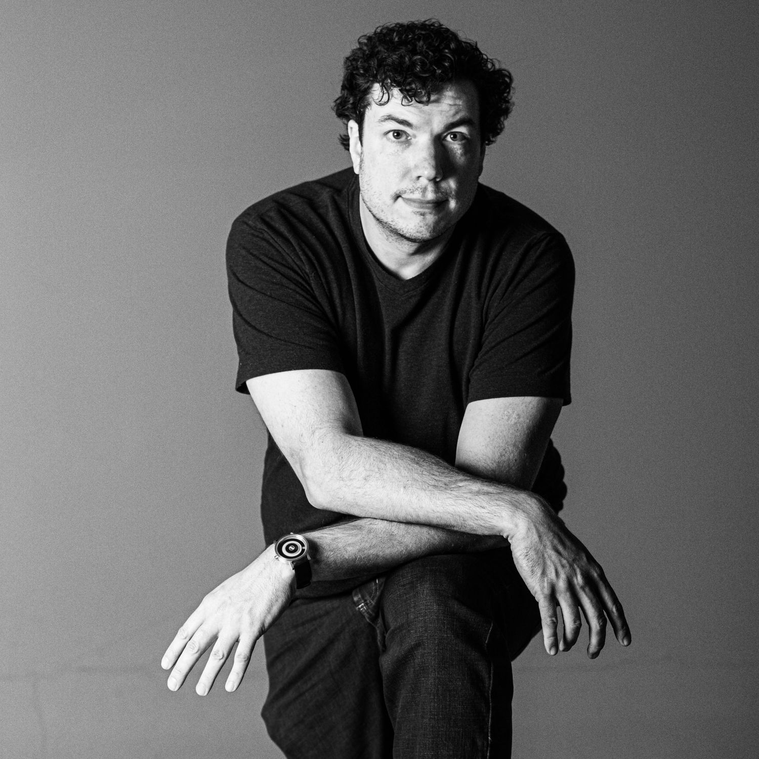 Black and white studio portrait of someone wearing a dark t-shirt seated with arms crossed.