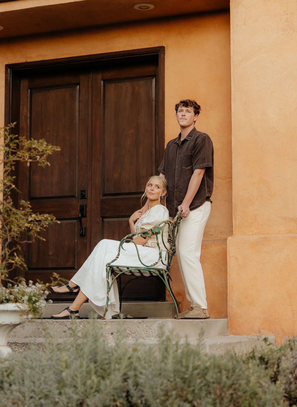 Couple posing on steps before rustic orange stucco home, woman seated in green chair, man standing beside her.