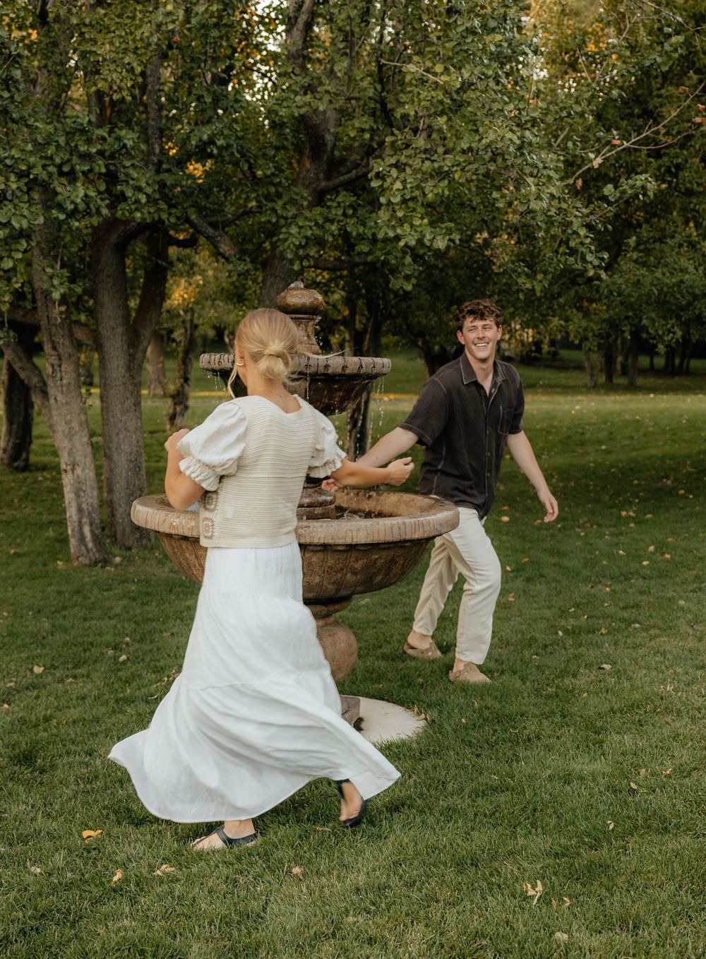 Playful couple chasing each other around a stone fountain in a lush green park setting.