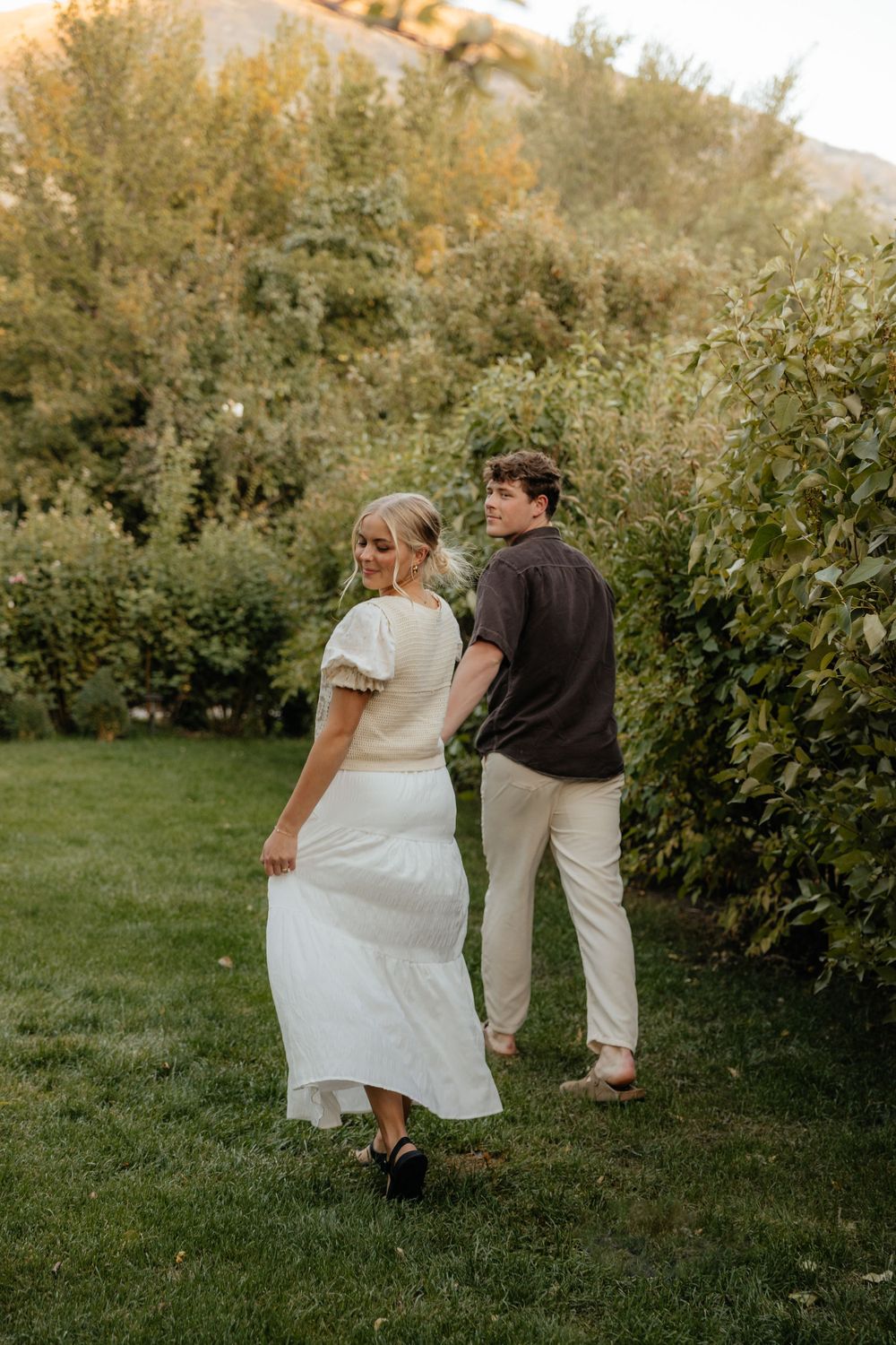 Couple walking hand in hand through a lush green garden pathway at sunset.