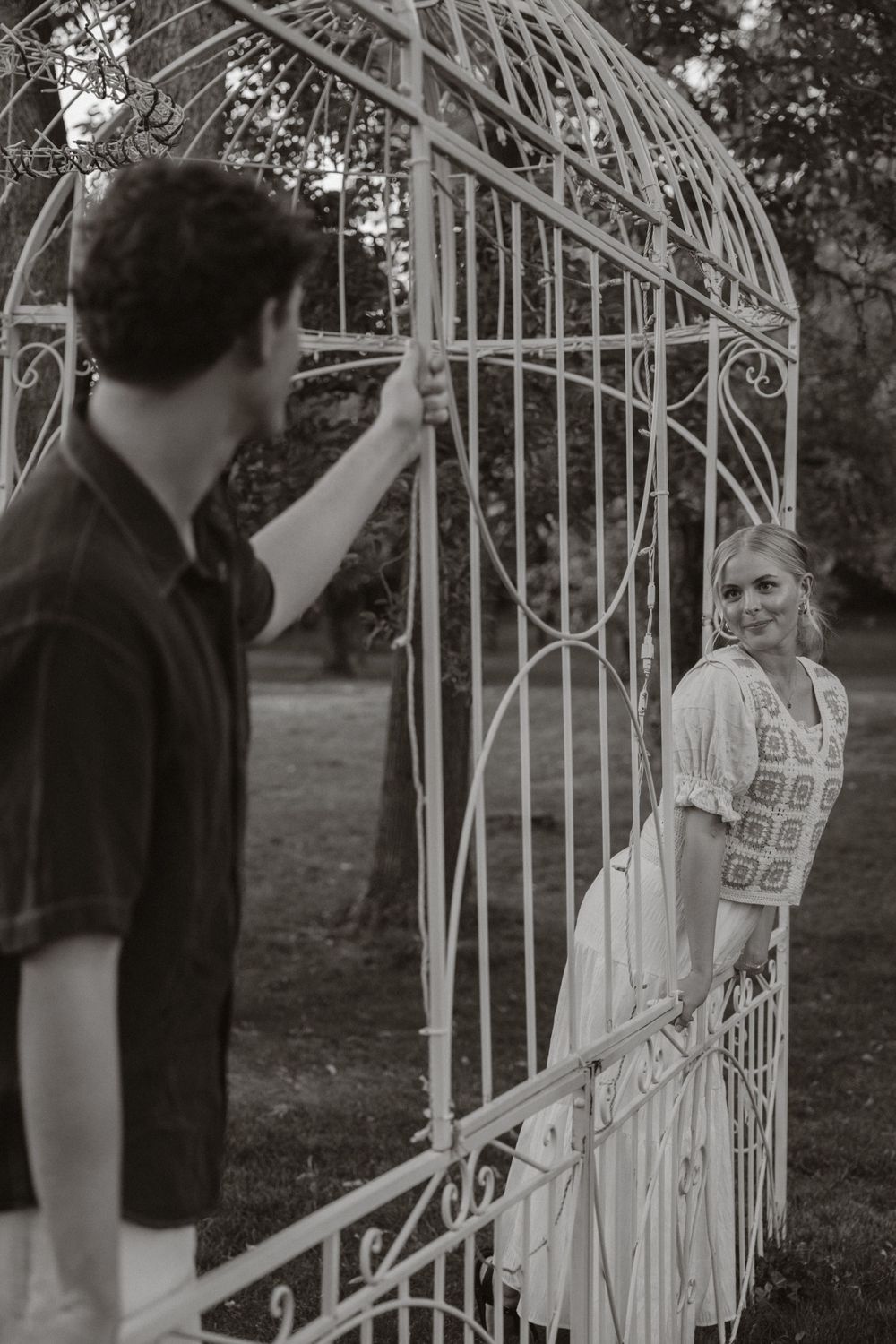 Black and white photo of man opening ornate white iron gate for smiling woman in garden.