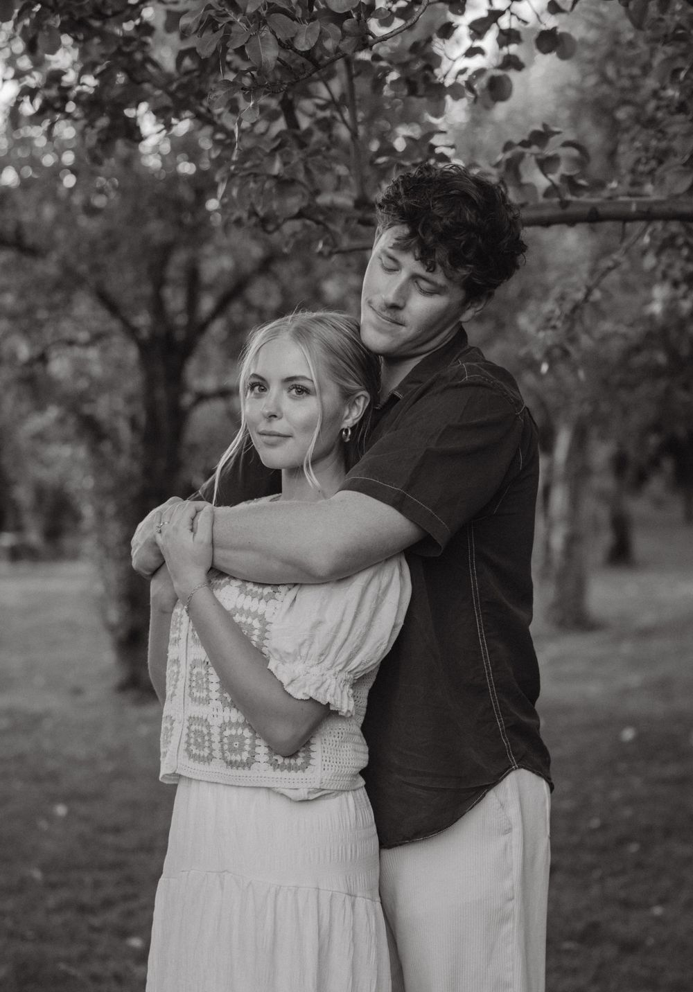 Black and white portrait of man tenderly embracing woman from behind under autumn orchard trees.