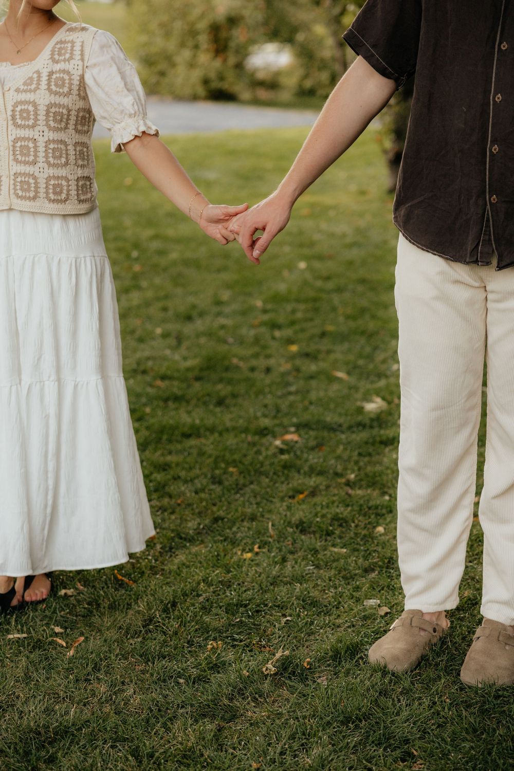 Close-up of couple holding hands gently on a green lawn, wearing neutral toned outfits.