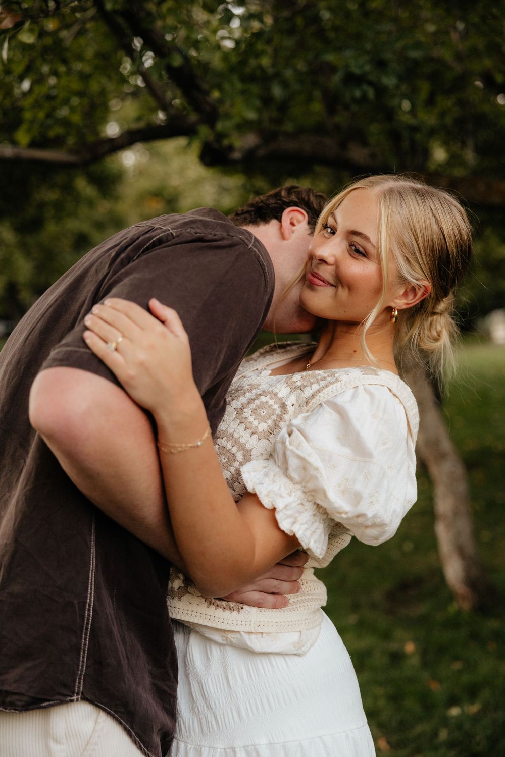 Man hugging woman from behind and kissing her cheek as she smiles under garden trees.