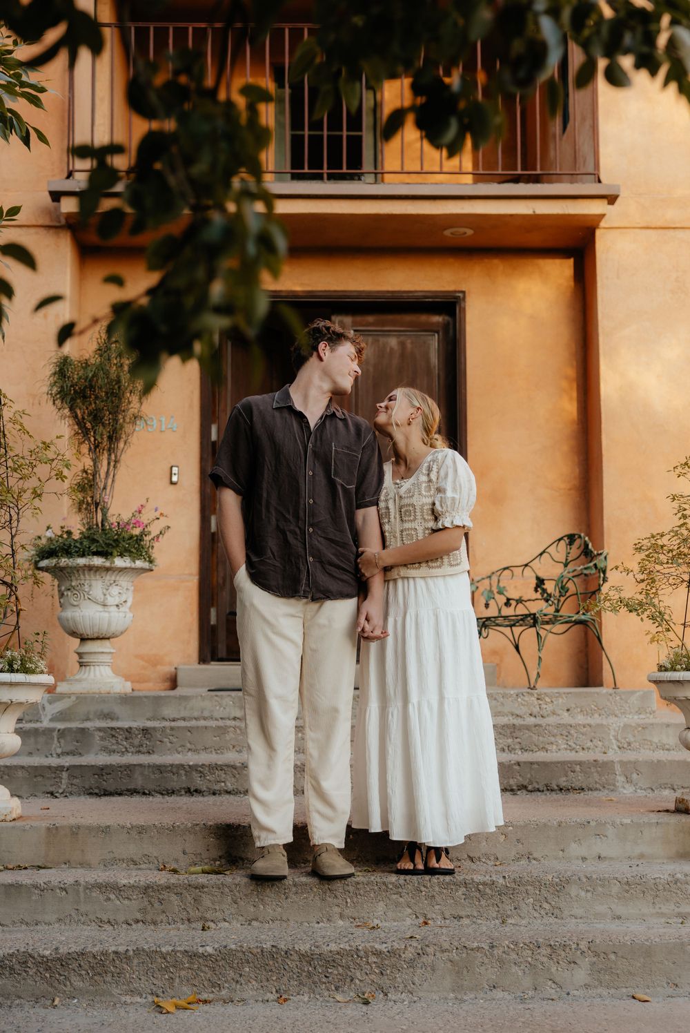 Couple gazing lovingly at each other on steps of a warm golden stucco Mediterranean building.