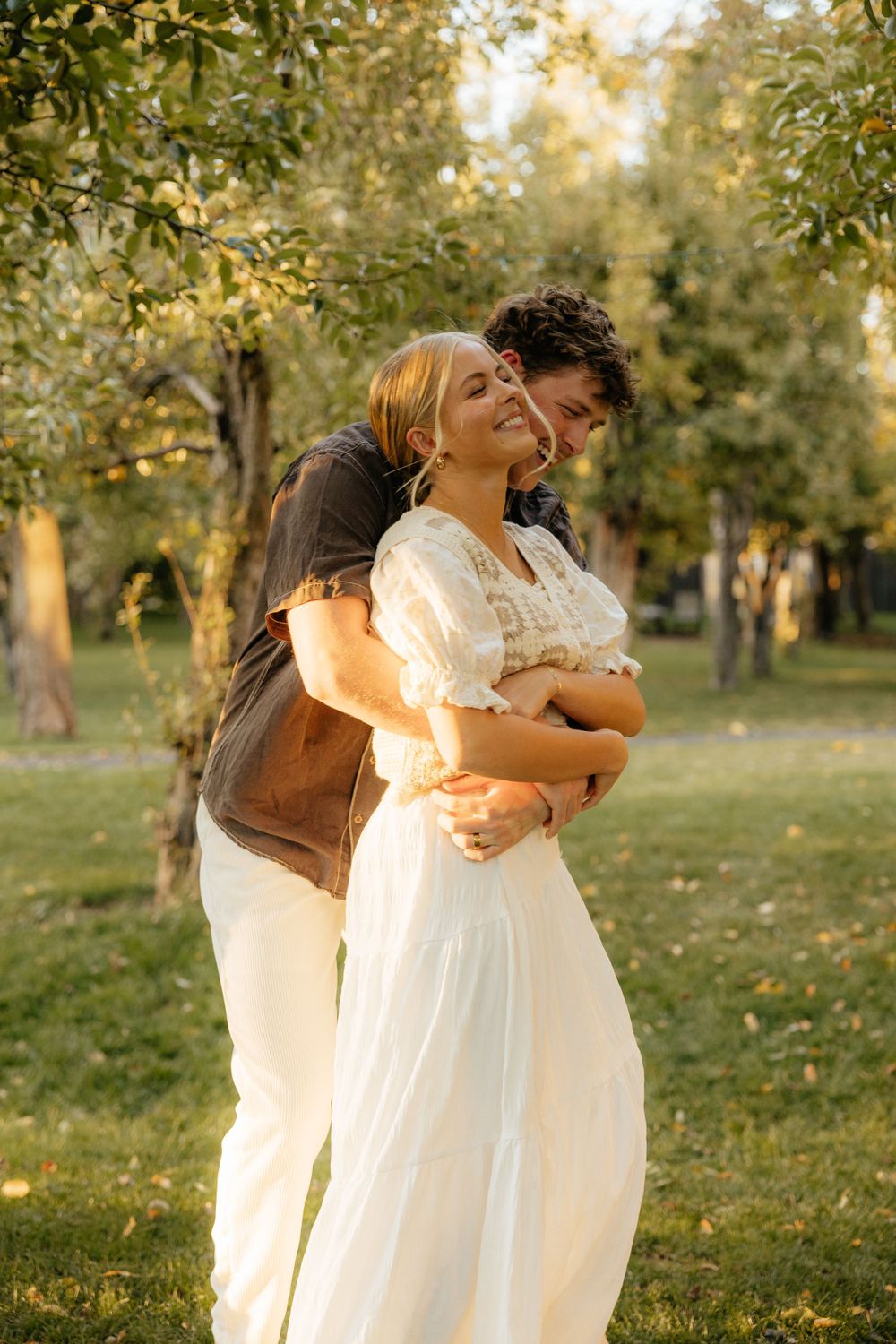 Man embracing woman in white dress from behind in a sunlit green park.
