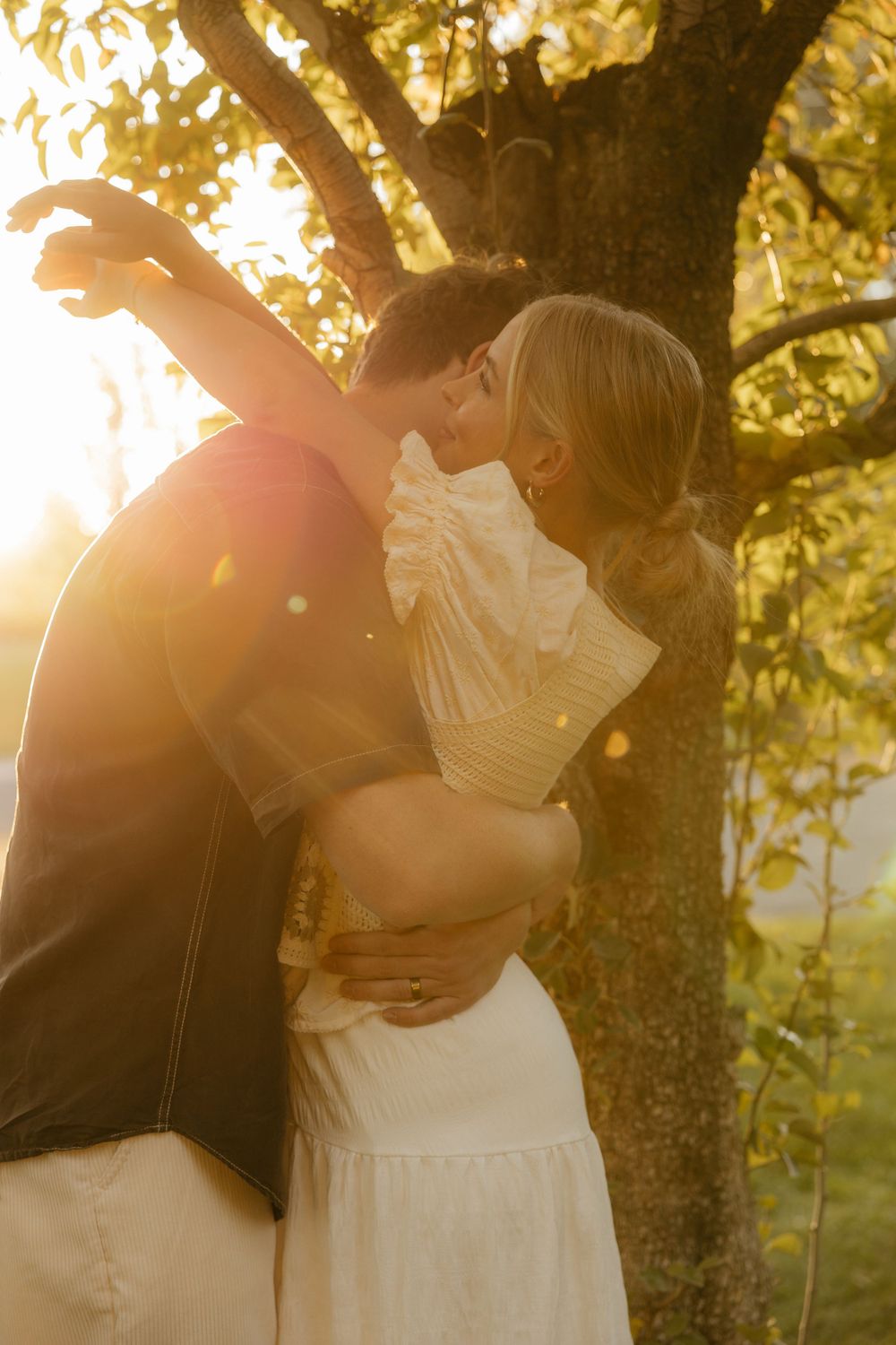 Couple sharing a romantic kiss bathed in warm golden sunset light by a tree.