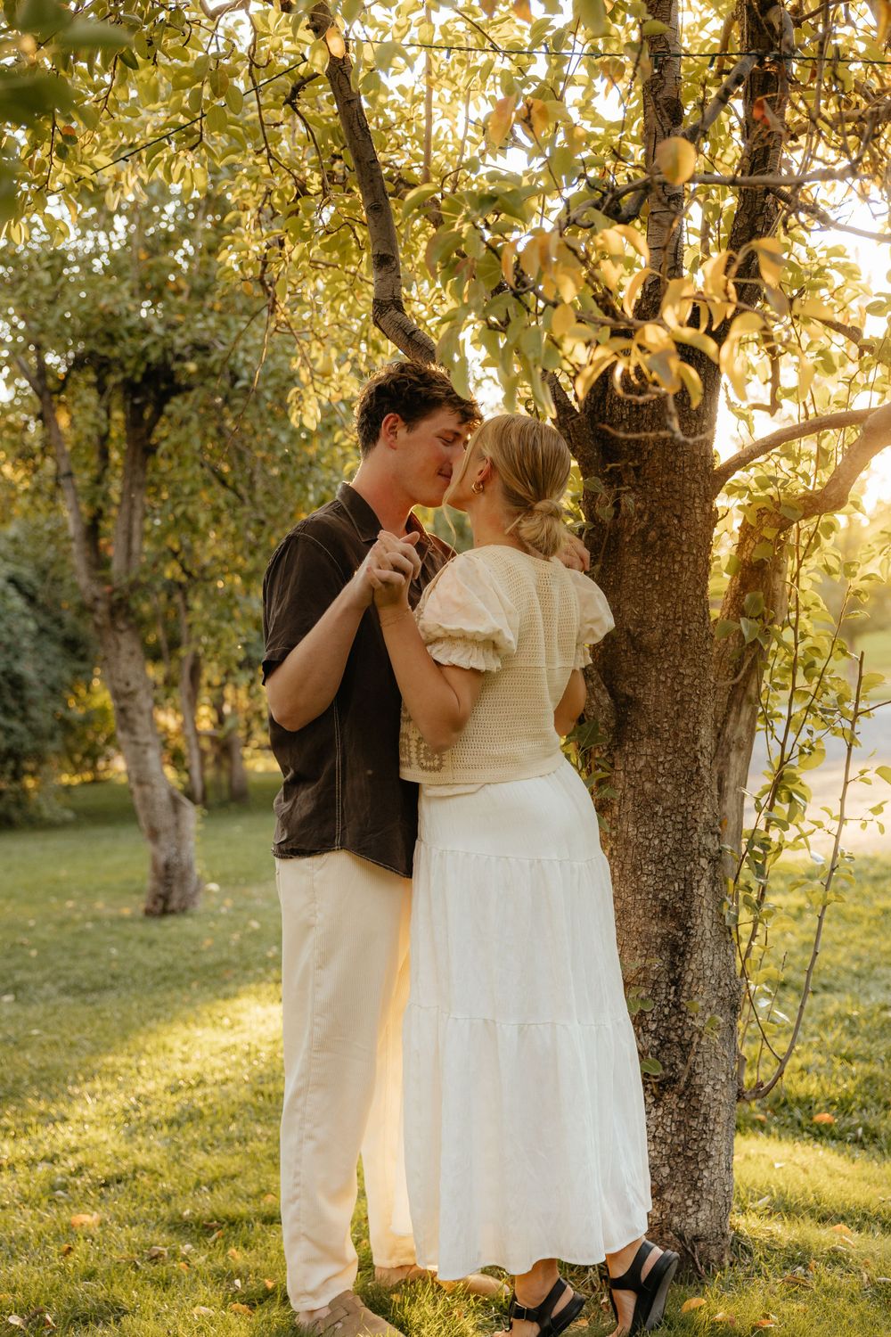 Couple sharing a tender kiss beneath a sunlit tree in a lush garden.