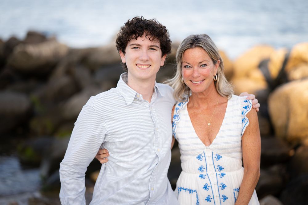 Two smiling people pose together on a rocky beach, wearing white outfits with ocean waves in the background.