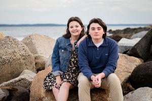 Two young people sitting on rocky shoreline with ocean view, wearing casual jackets in an outdoor portrait.