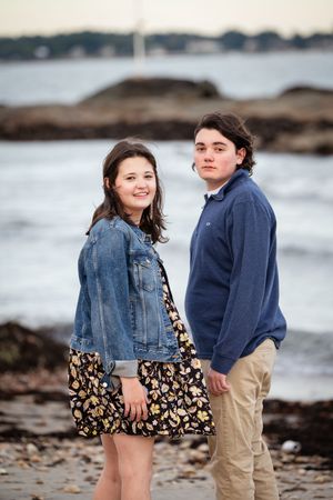 Two teenagers pose together on a rocky beach, girl in denim jacket and floral dress, boy in blue sweater and khakis.
