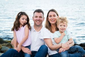 A happy family sits together on rocks near the ocean in coordinated blue and white outfits during a beach photoshoot.