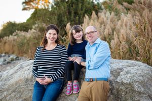 Family of 3 sit together on a large boulder with tall ornamental grasses behind them on an autumn afternoon.