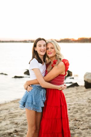 Mother and daughter embrace and laugh together on a beach at sunset while wearing casual summer outfits.