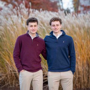 Two young men in matching quarter-zip sweaters and khaki pants pose together in front of tall ornamental grasses.