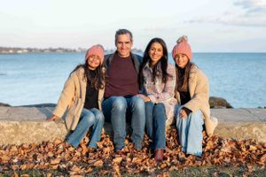 A group of four people wearing casual winter clothing sit together on fallen leaves by the ocean during sunset.