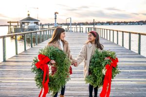 Two people stand on a wooden pier holding large green Christmas wreaths decorated with red bows at sunset.