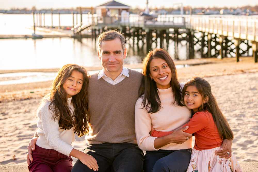 Family of four sitting together on a sandy beach with a wooden pier in the background during golden hour.