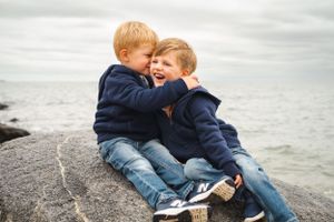 Two children in navy sweaters laugh together while sitting on rocky shoreline by the ocean.