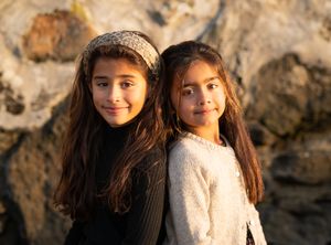 Two young sisters with long dark hair pose together outdoors at sunset against a rocky backdrop.