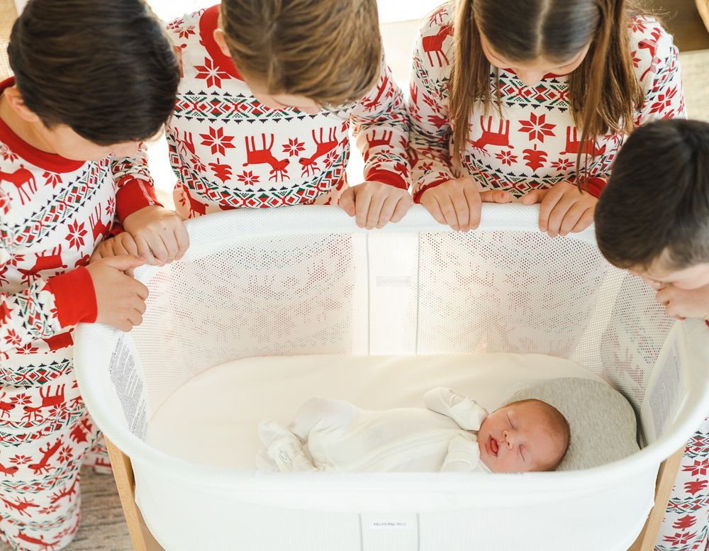 Four children in matching red and white patterned sleepwear peer down at a newborn baby lying in a white bassinet.