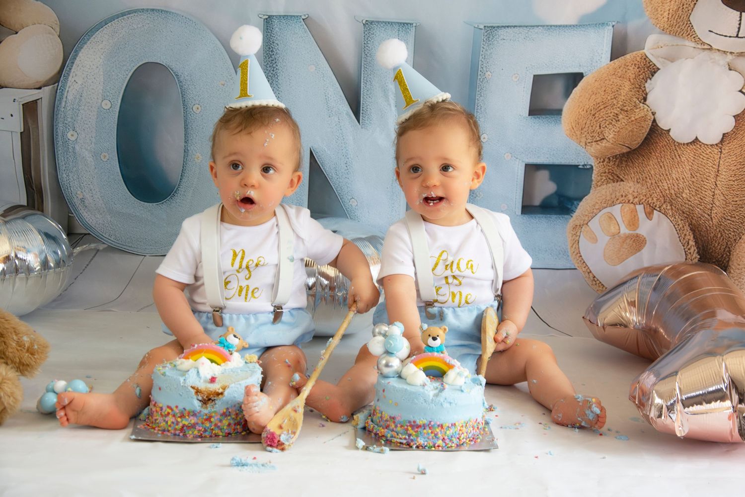 Two babies in birthday party hats celebrate their first birthday with matching blue cakes and stuffed animals.