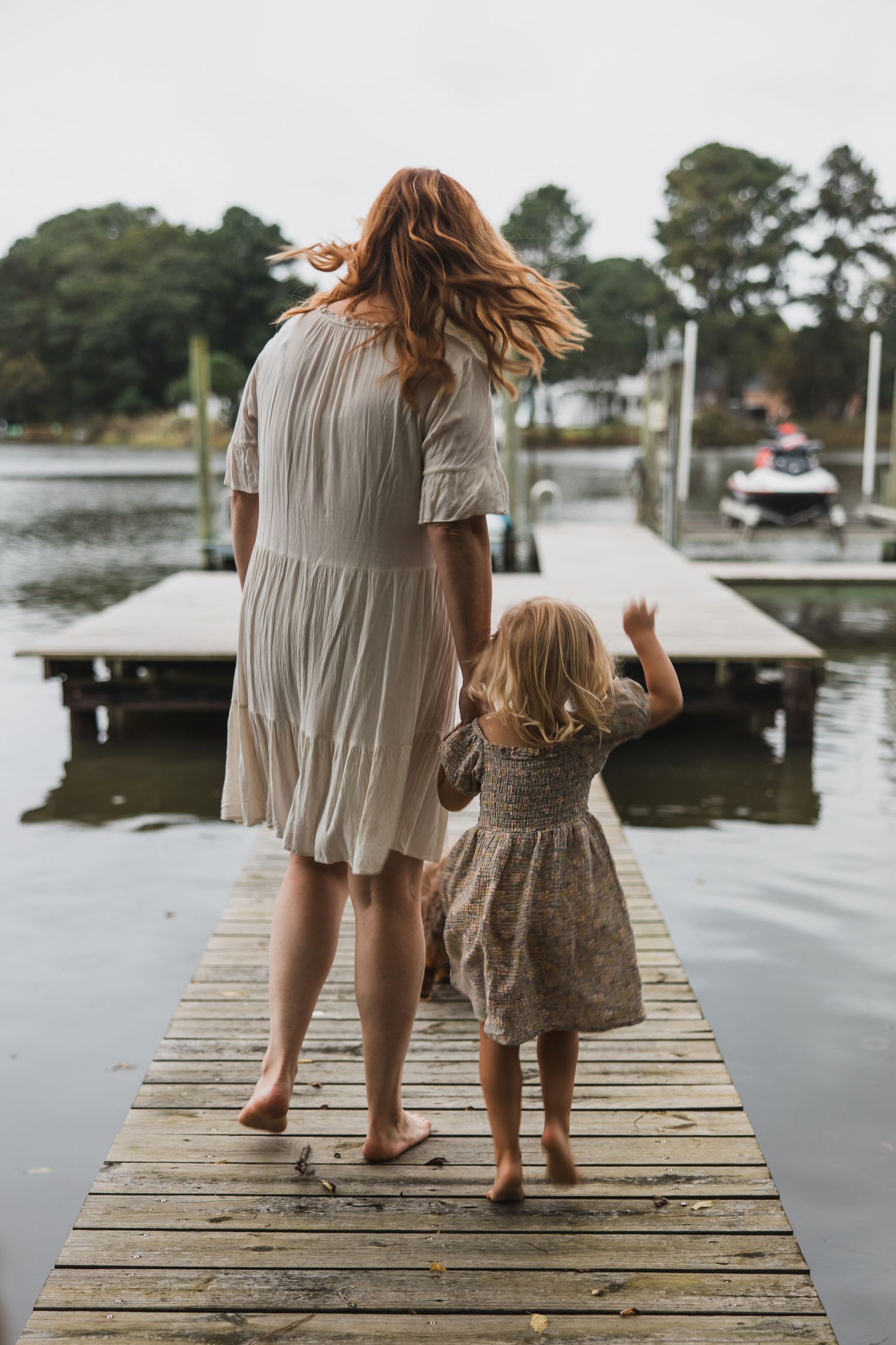 Mother and child walking barefoot on wooden dock by calm waters.