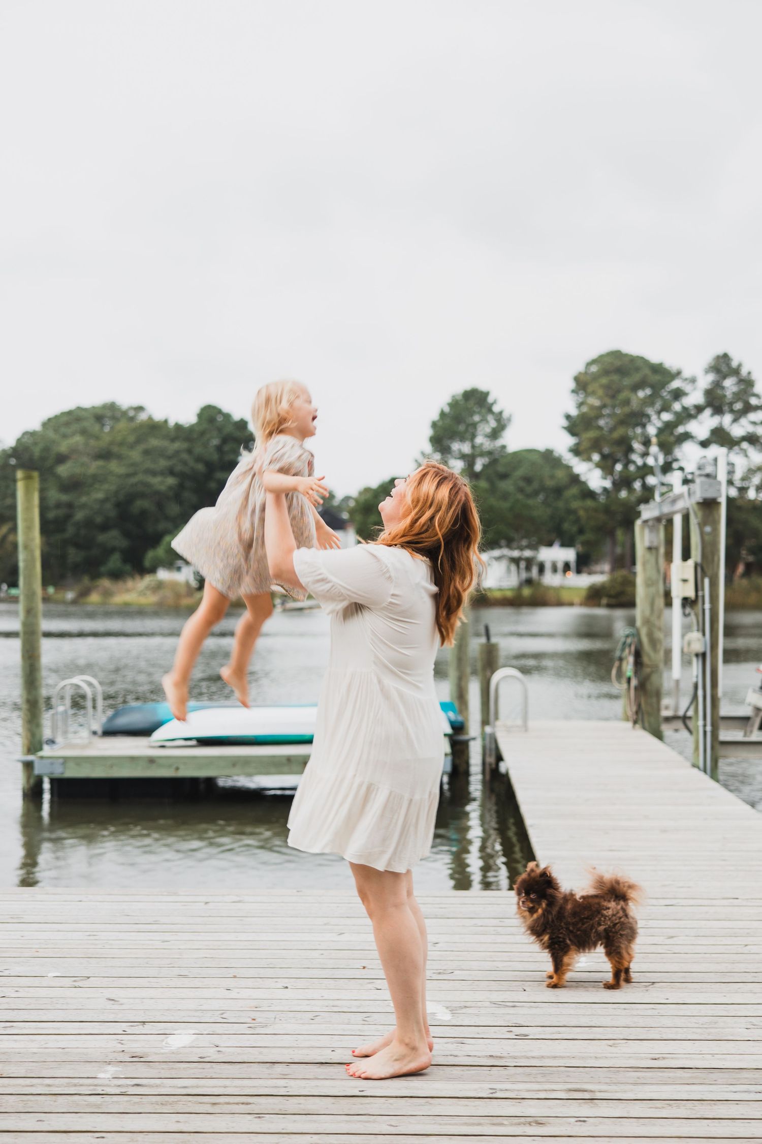 Parent lifting child playfully on wooden dock while small dog watches.