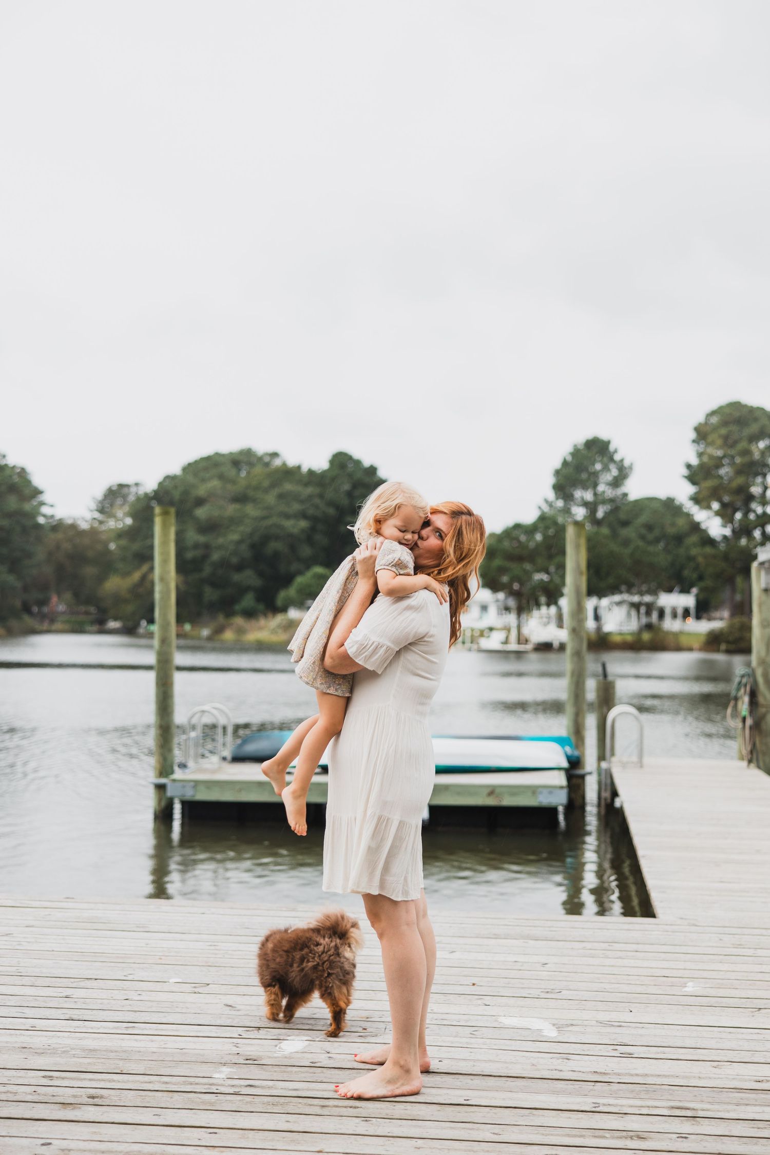 Parent embracing child on waterfront dock with small brown dog nearby.