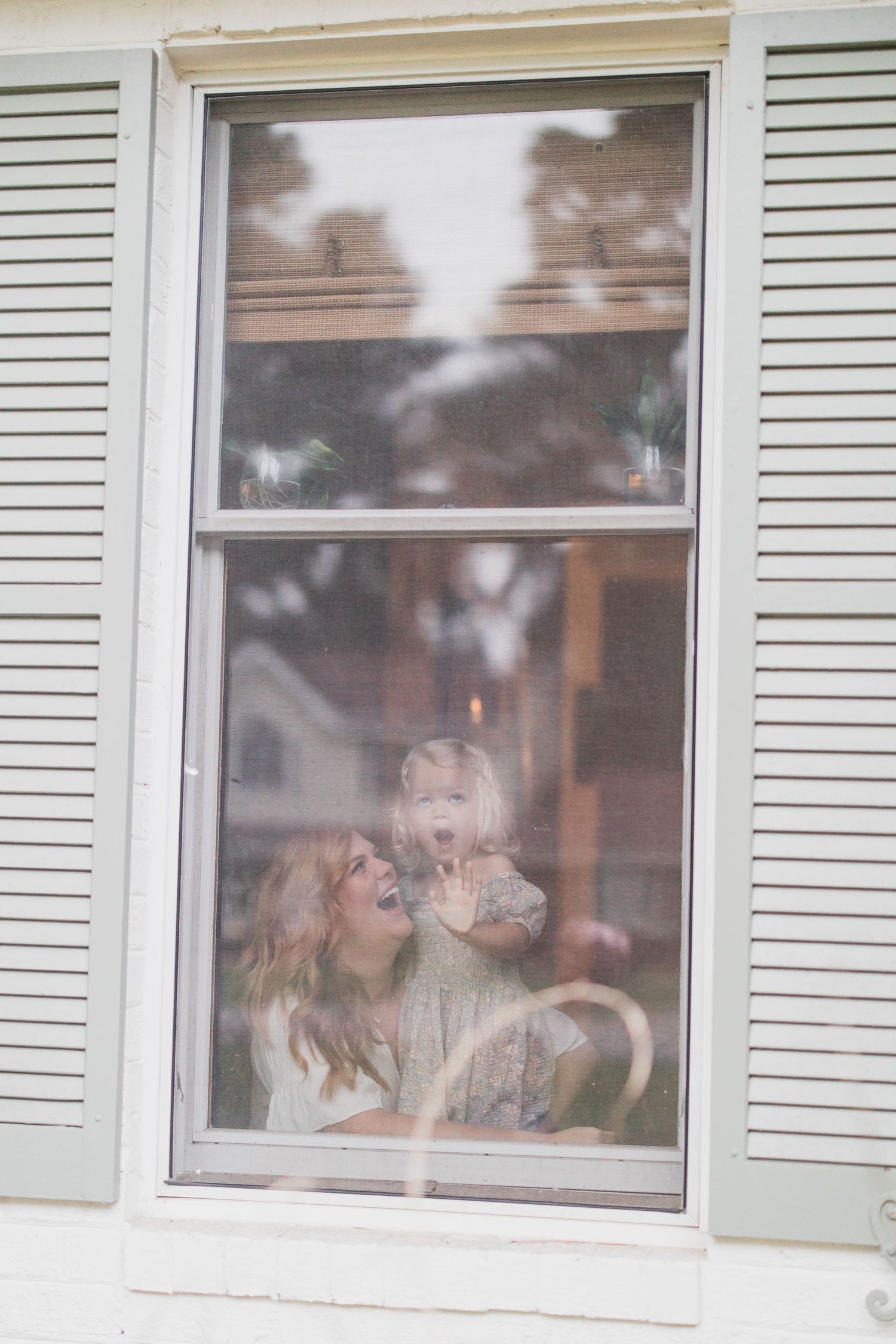 Candid moment captured through window with white shutters showing joyful interaction.