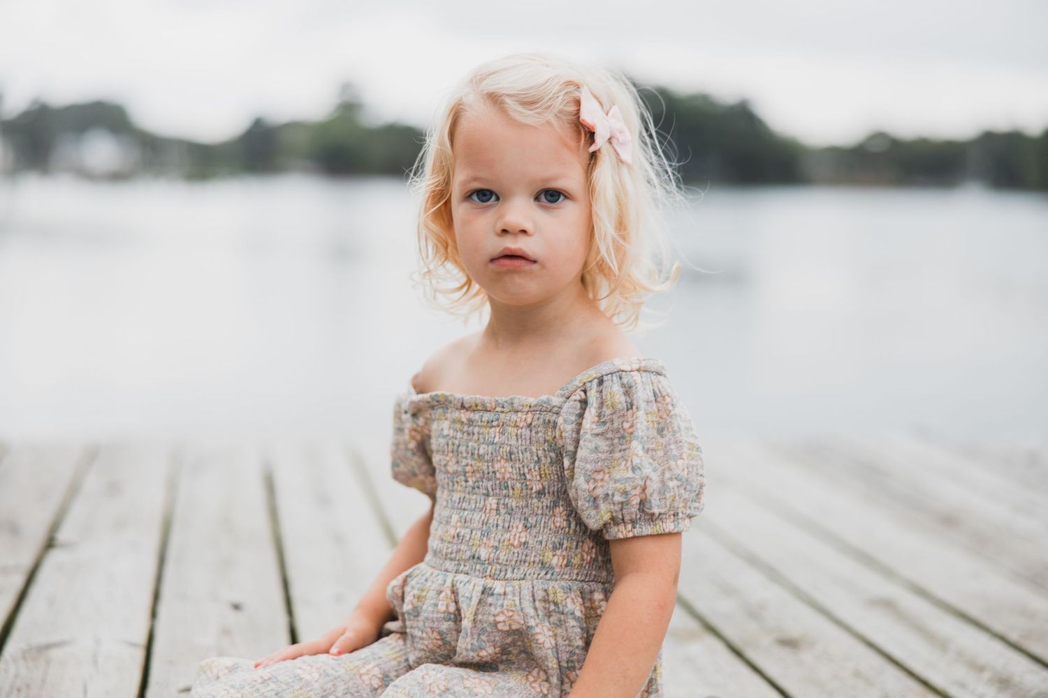 Small child in vintage-style dress sitting on wooden dock by water.