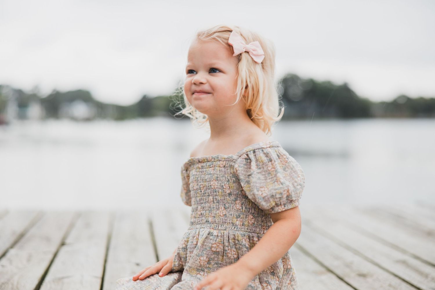 Young child in floral dress smiling while sitting on lakeside dock.