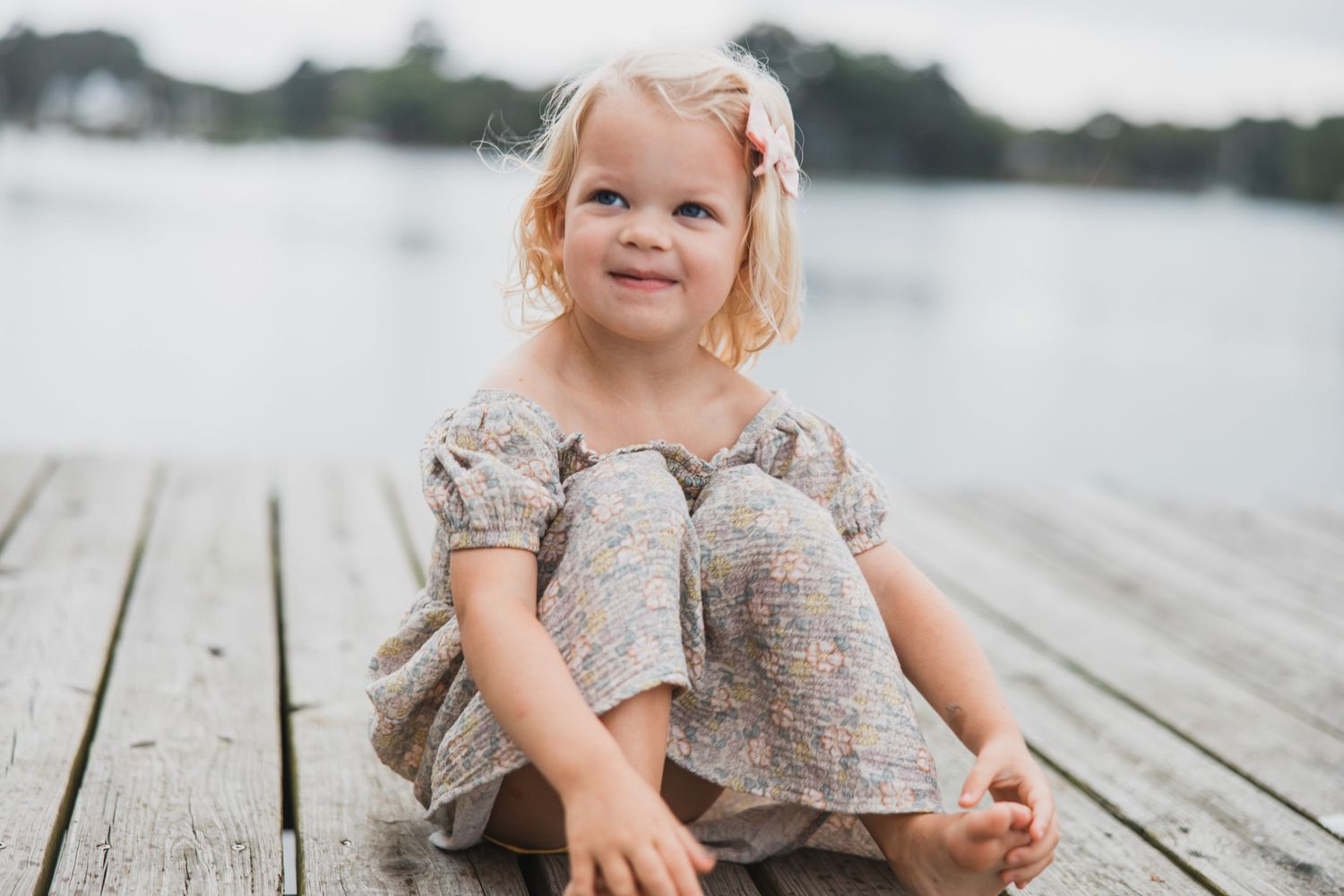 Child in vintage dress sitting cross-legged on wooden dock near water.