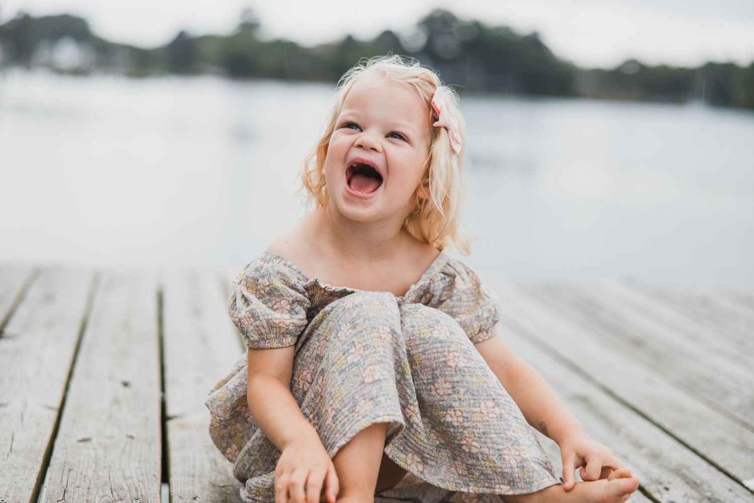 Joyful child laughing while sitting on wooden dock wearing floral dress.