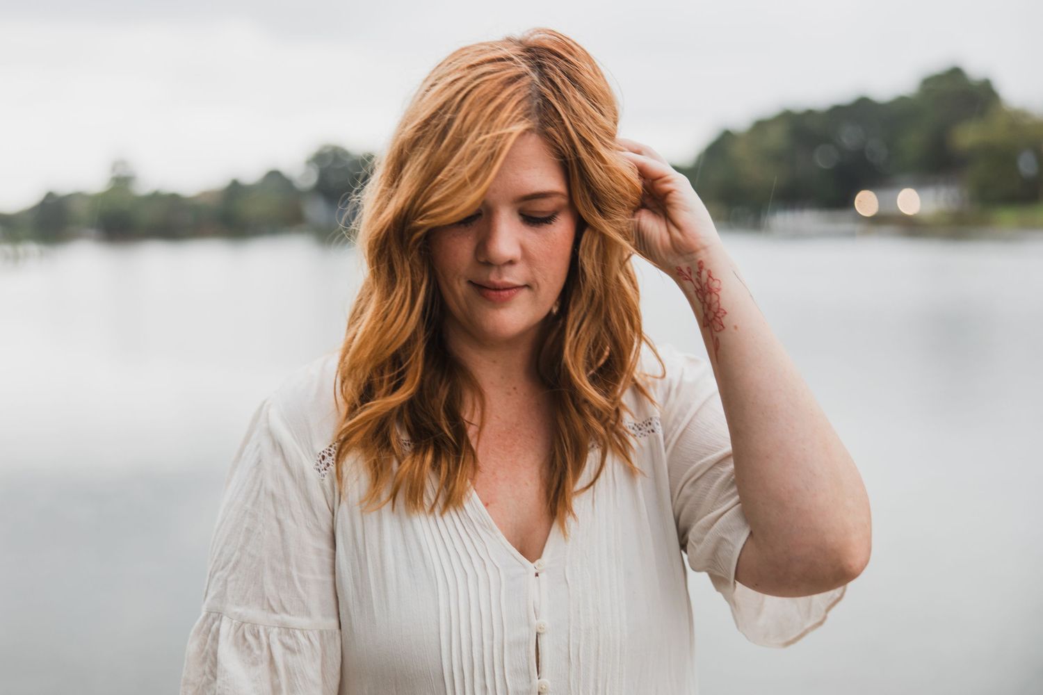 Woman in white blouse with wavy hair looking down thoughtfully by lake.