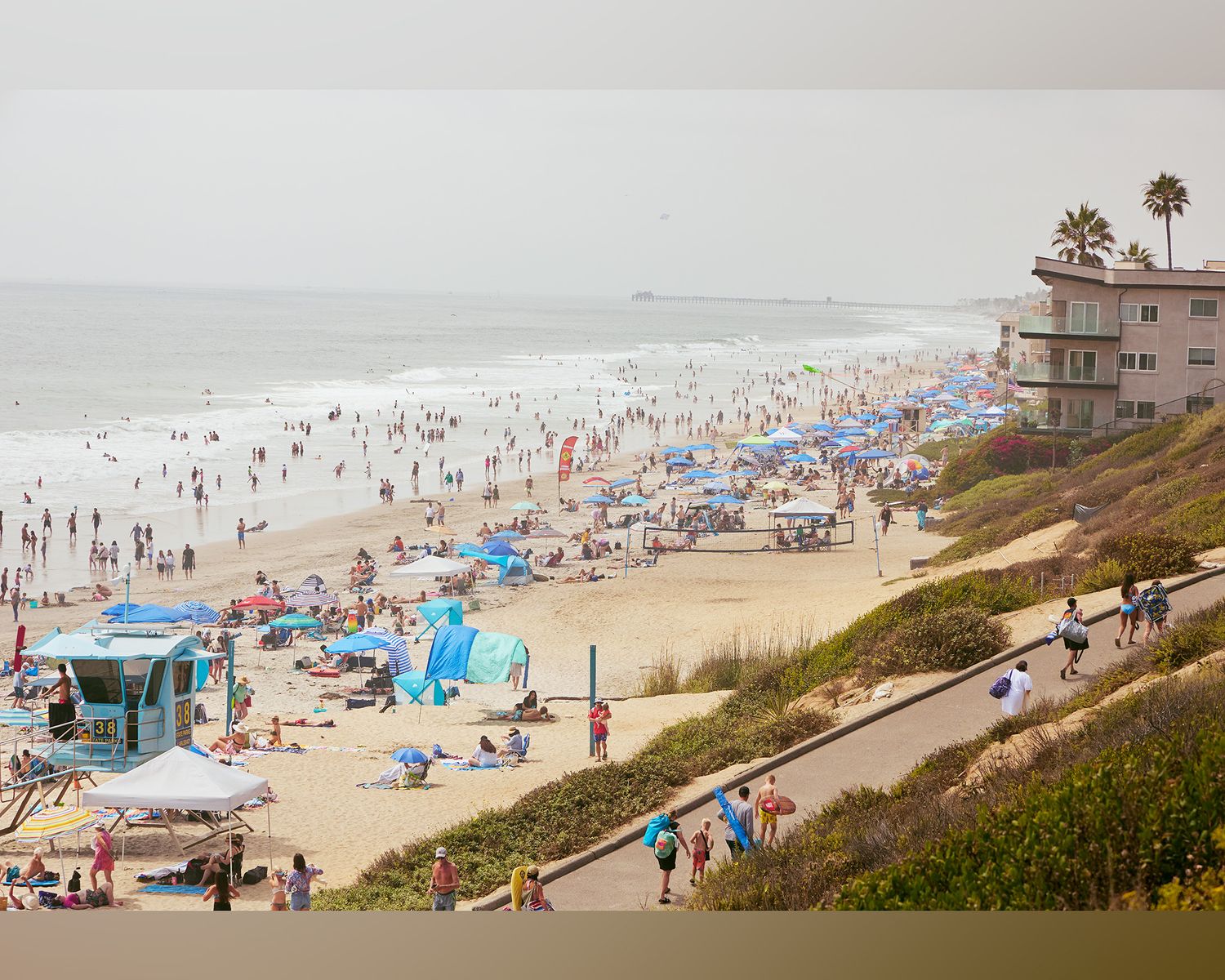 Panoramic view of a crowded California beach with colorful umbrellas and beachgoers dotting the sandy shoreline on a sunny day.