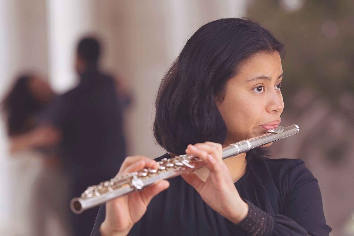 A musician plays the flute while wearing a black sweater in a softly lit indoor setting.