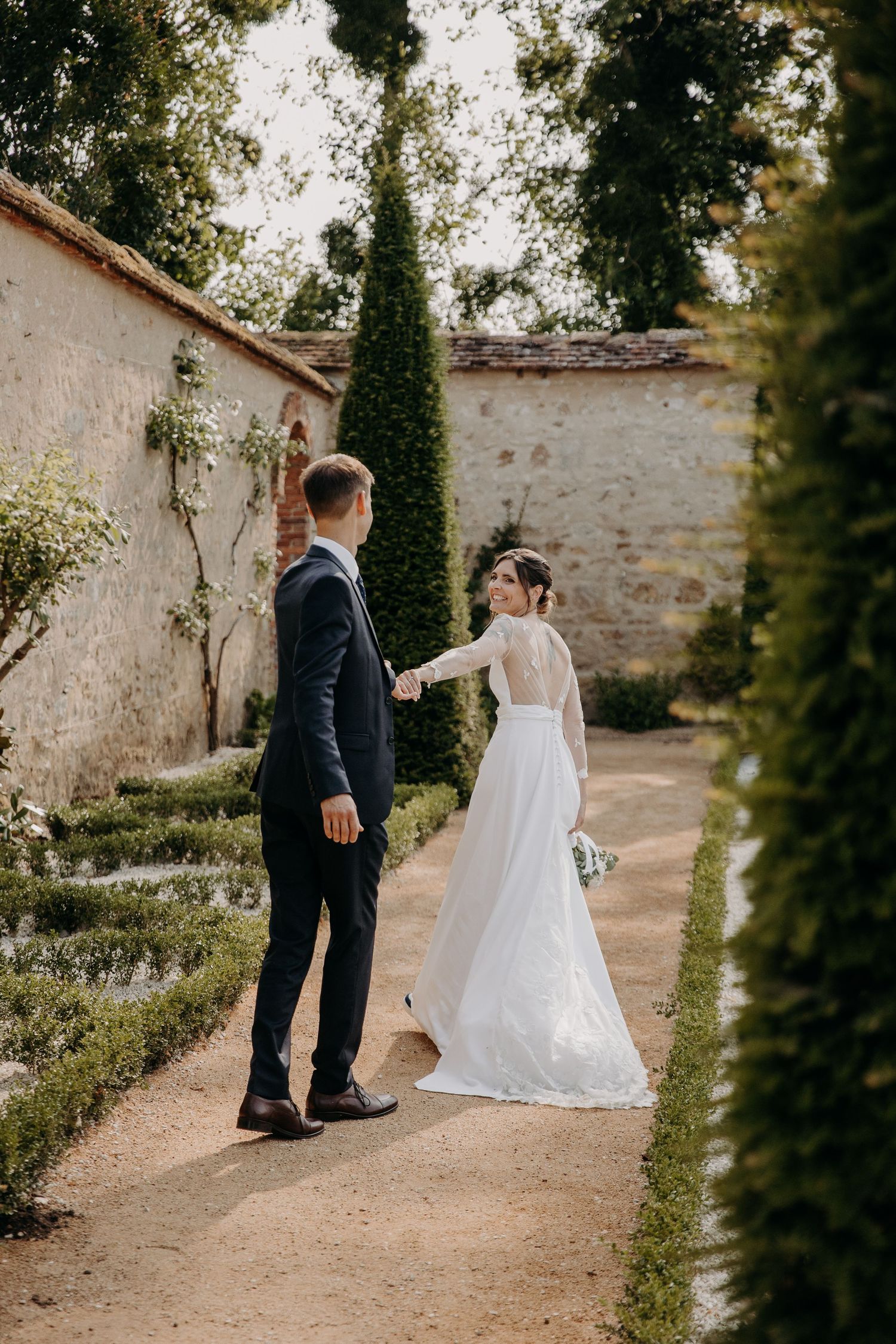 Newlyweds walking along garden path surrounded by stone walls and greenery.