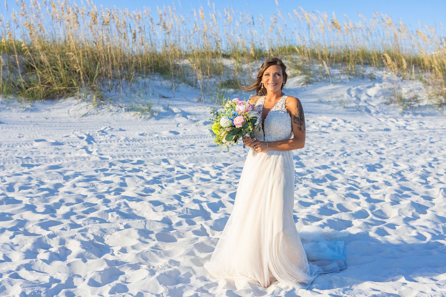 A bride holding a bouquet stands in white sand dunes at the beach.