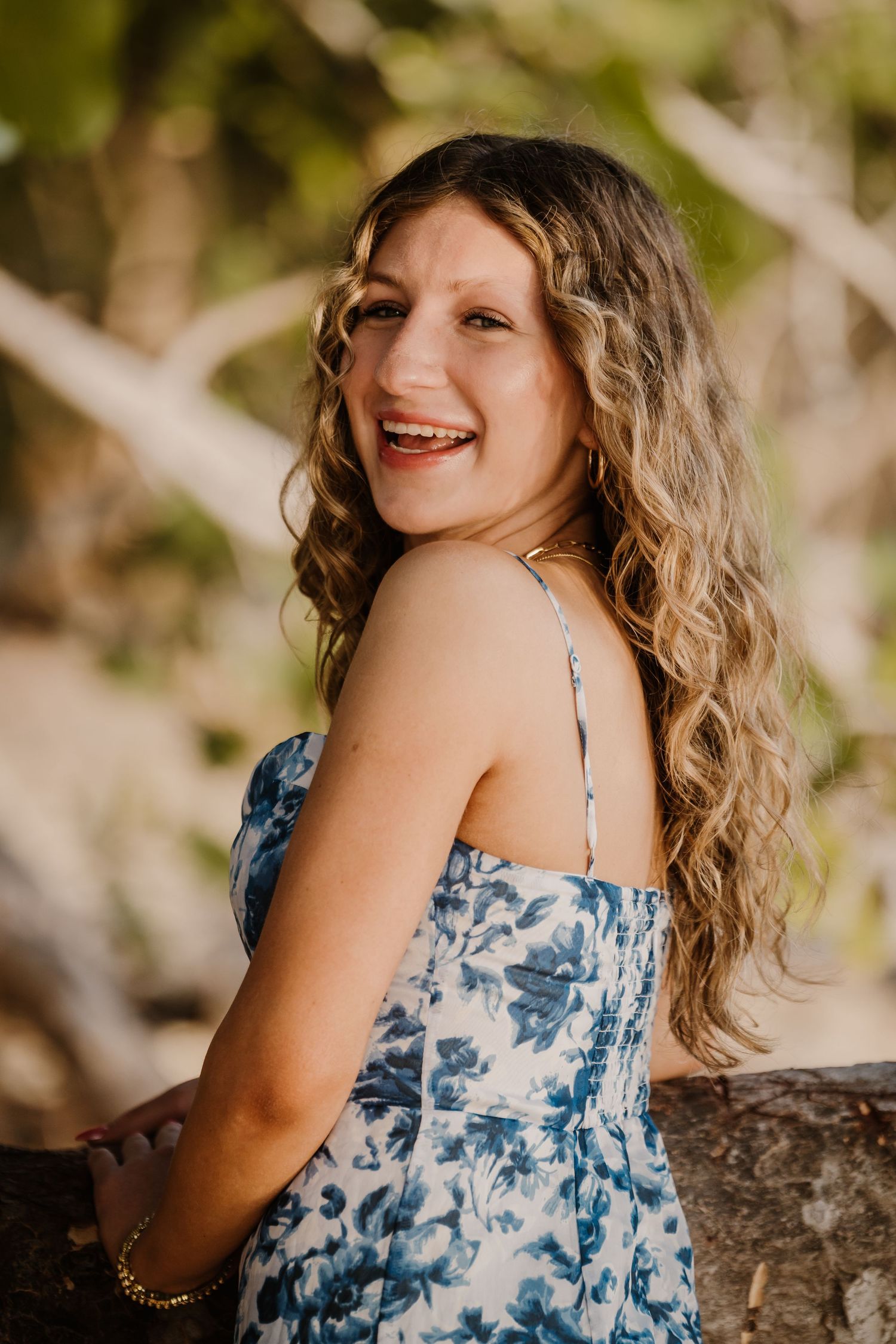 A person in a blue and white floral sundress smiles while posing outdoors with wavy blonde hair in natural lighting.