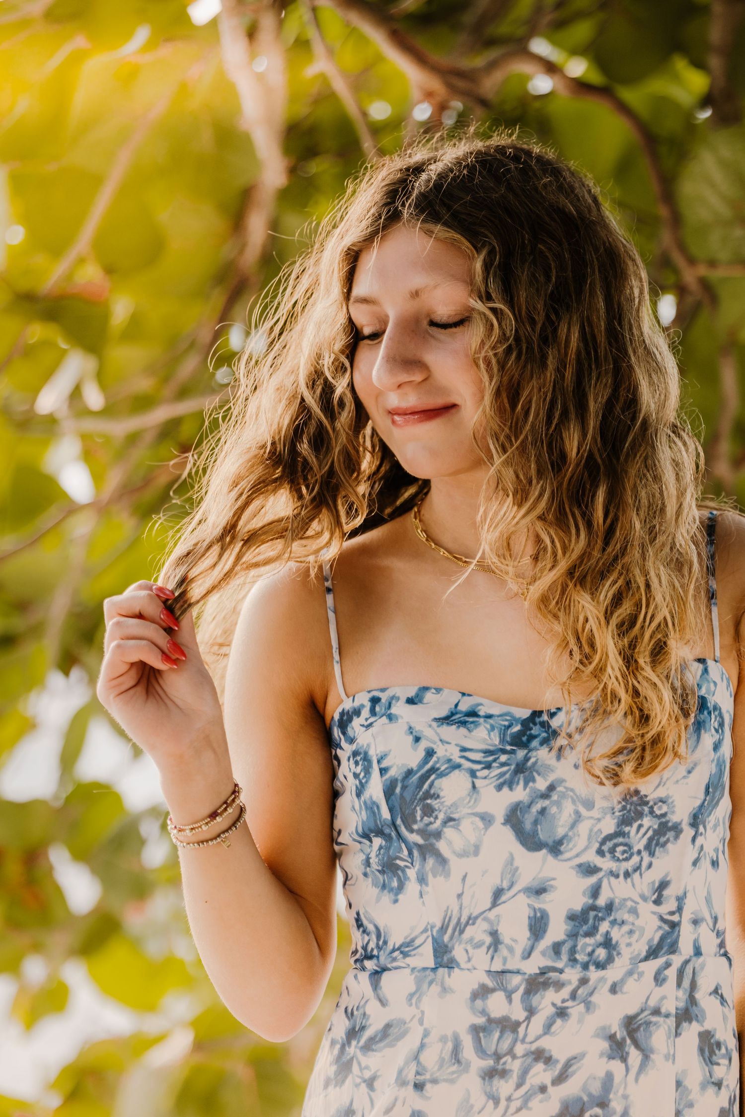 A person in a blue and white floral dress twirls among sunlit leaves with wavy hair and a peaceful expression.