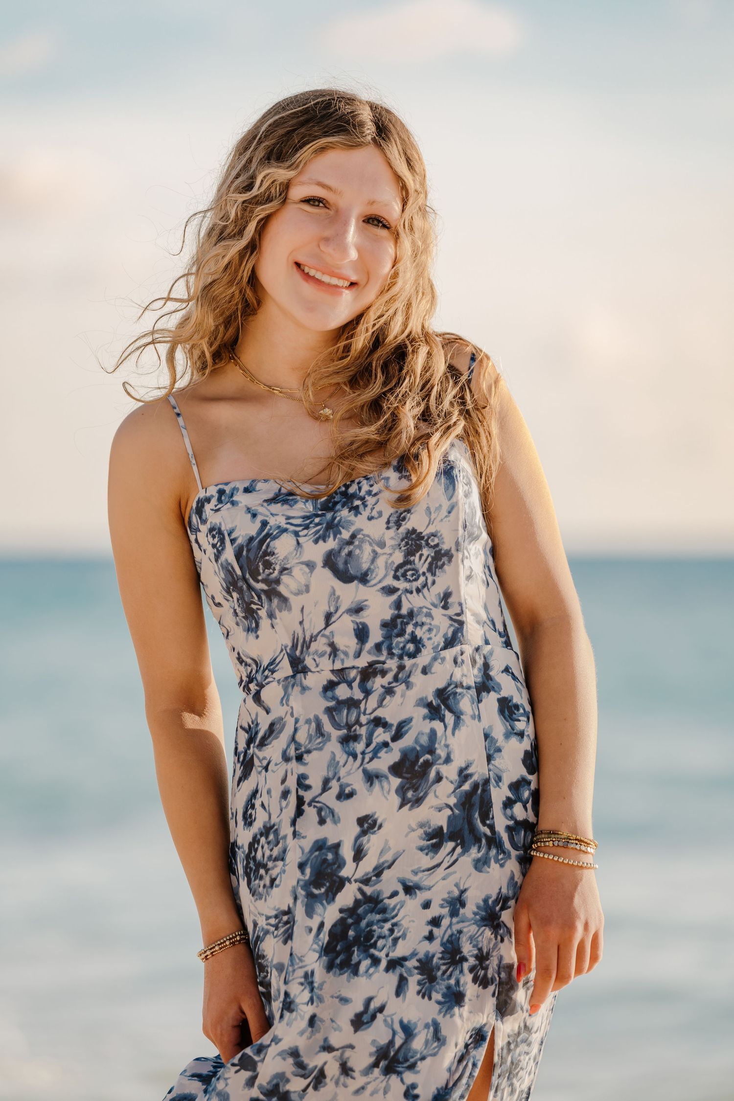 A person in a blue and white floral sundress poses on a beach with soft clouds and ocean in the background.