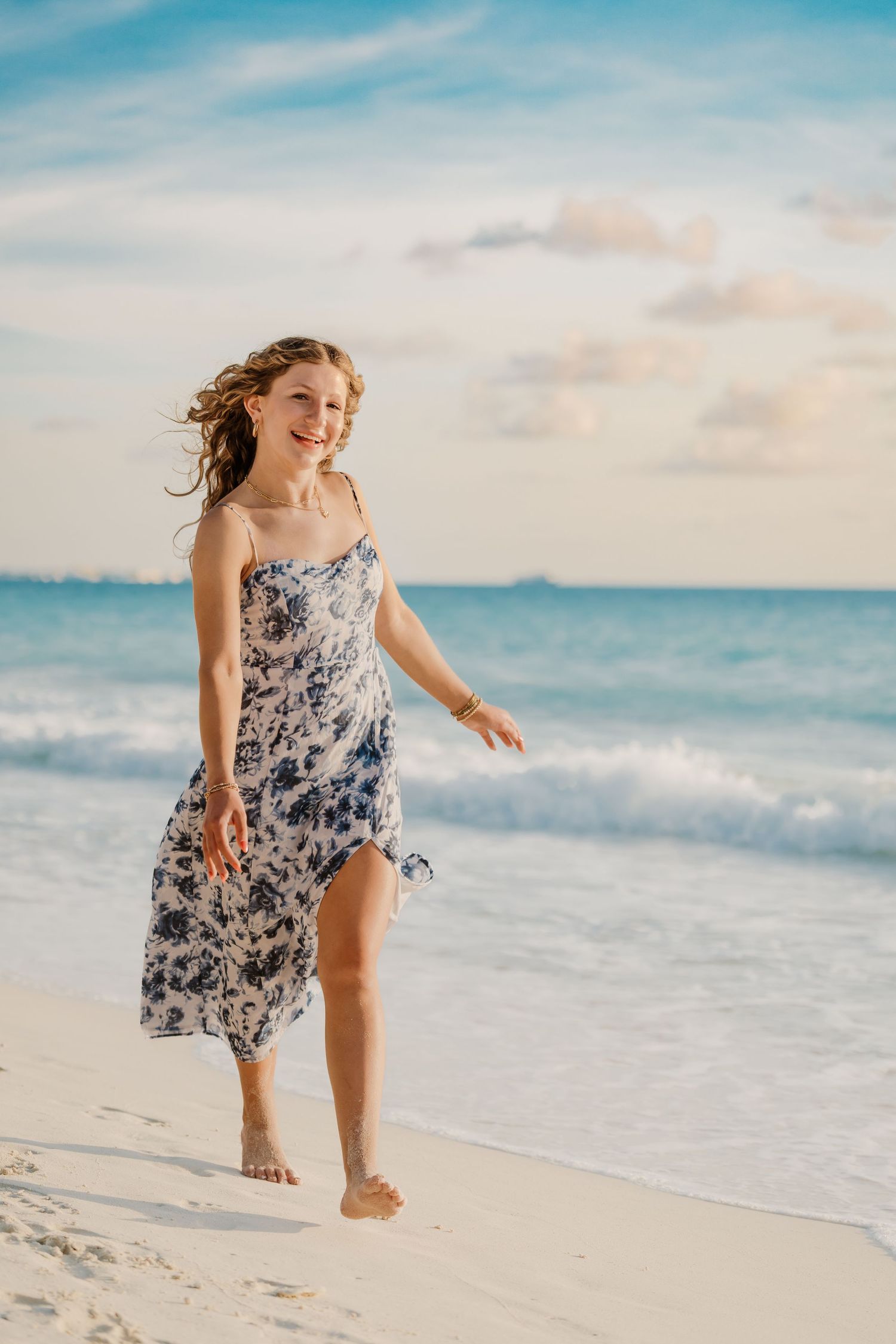 A feminine figure in a floral print dress walks joyfully along a tropical beach with waves and blue sky in the background. Senior Photos